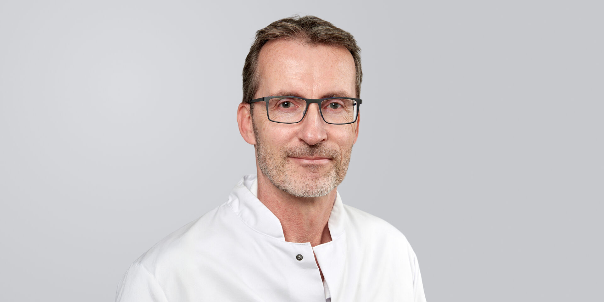 Mann mit Brille, Bart und weißem Kittel vor einem grauen Hintergrund.