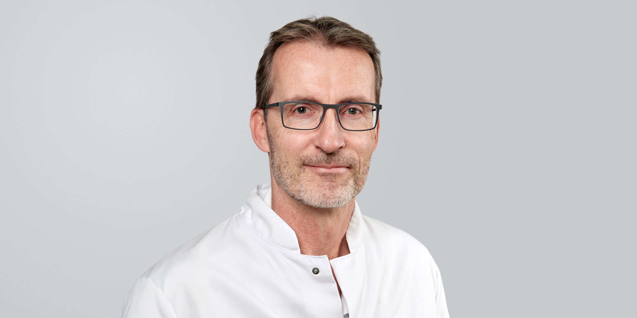 Mann mit Brille, Bart und weißem Kittel vor einem grauen Hintergrund.