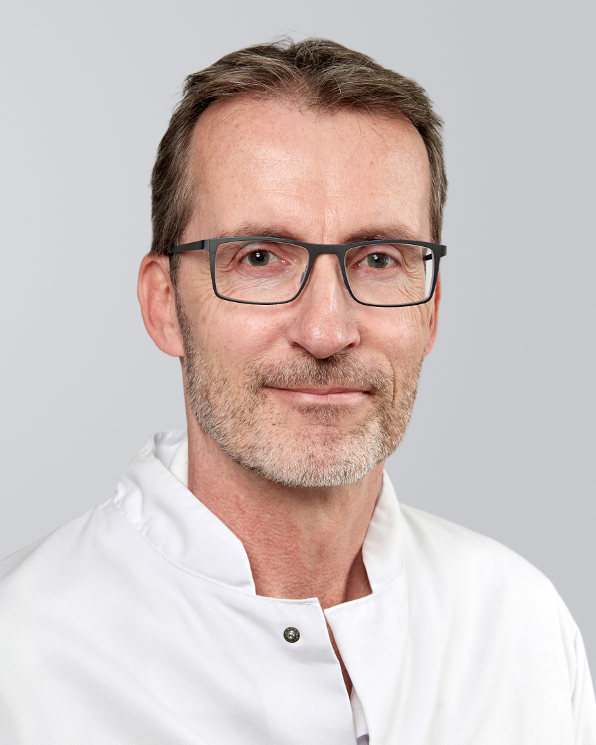 Mann mit Brille, Bart und weißem Kittel vor einem grauen Hintergrund.