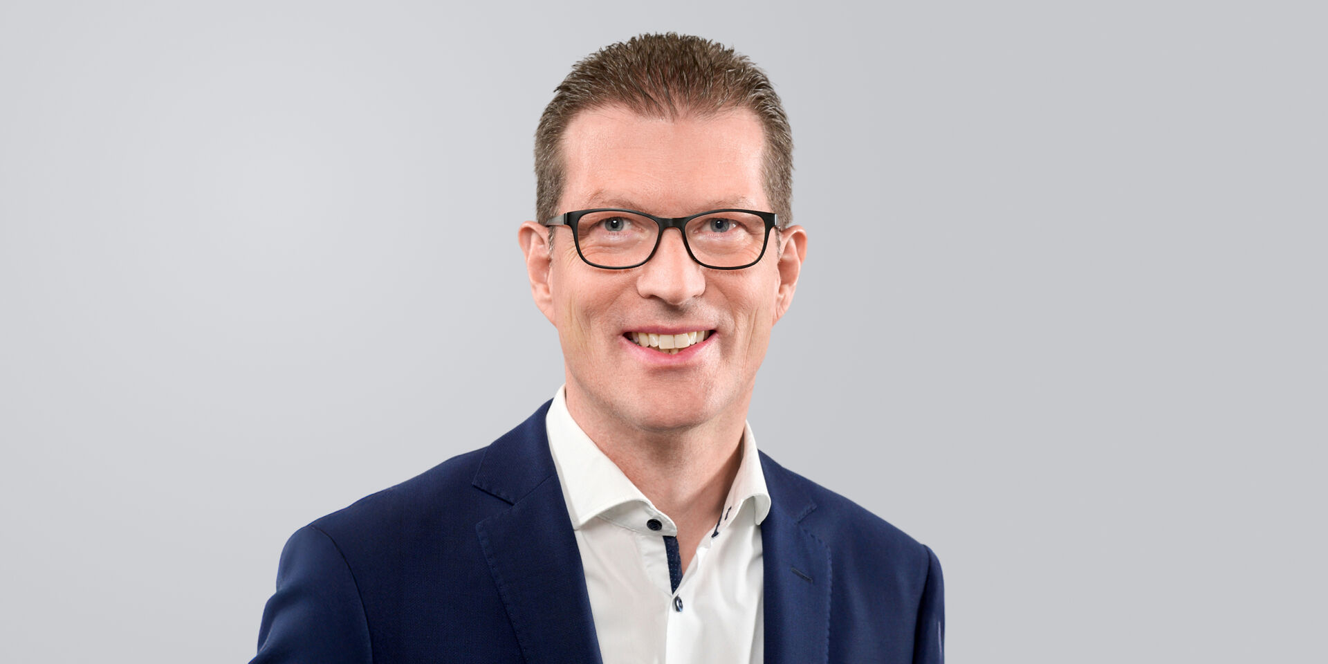 Mann mit Brille, kurzem Haar und dunkelblauem Anzug vor einem grauen Hintergrund.