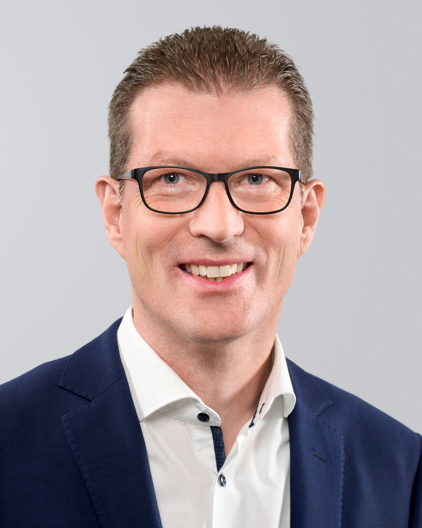 Mann mit Brille, kurzem Haar und dunkelblauem Anzug vor einem grauen Hintergrund.