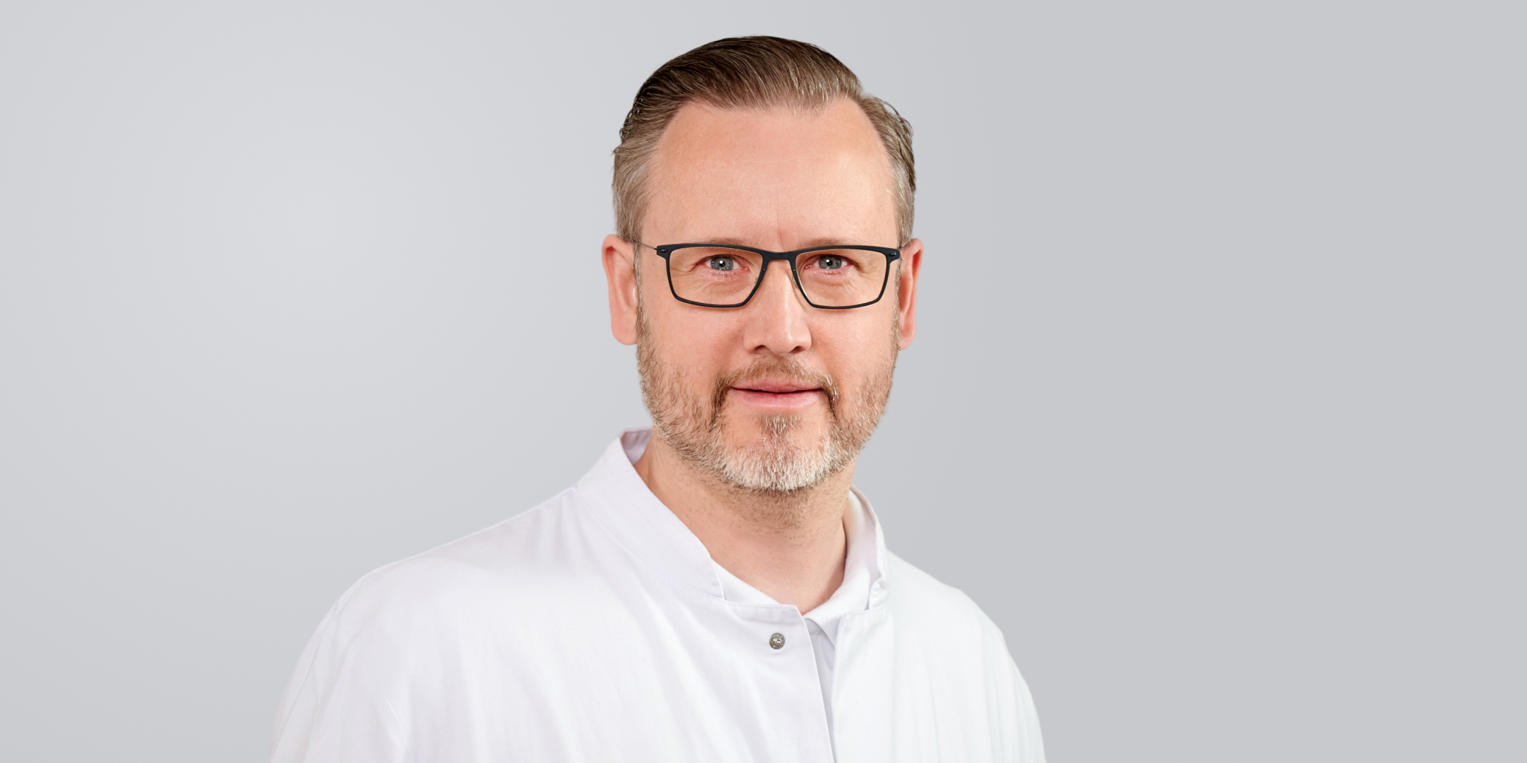 Mann mit Brille, grauem Bart und weißem Hemd vor einem grauen Hintergrund.