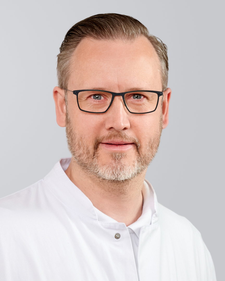 Mann mit Brille, grauem Bart und weißem Hemd vor einem grauen Hintergrund.