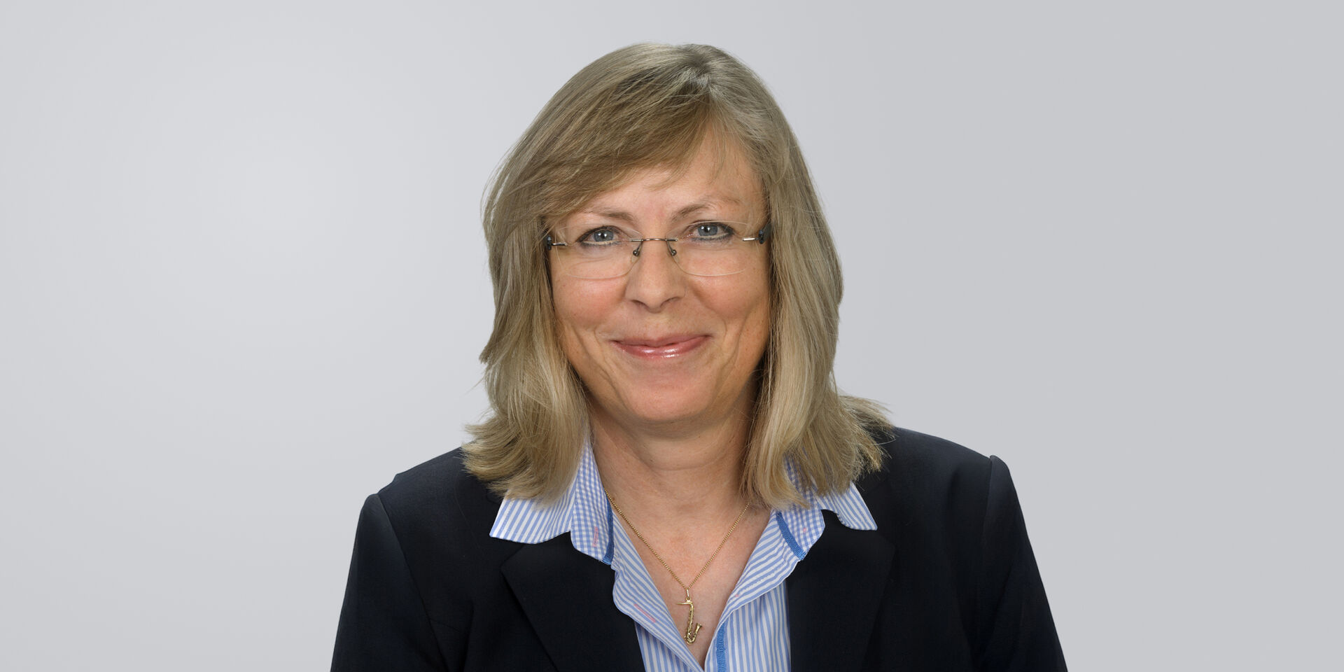 Frau mit blonden, schulterlangen Haaren, Brille und dunklem Blazer vor neutralem Hintergrund.