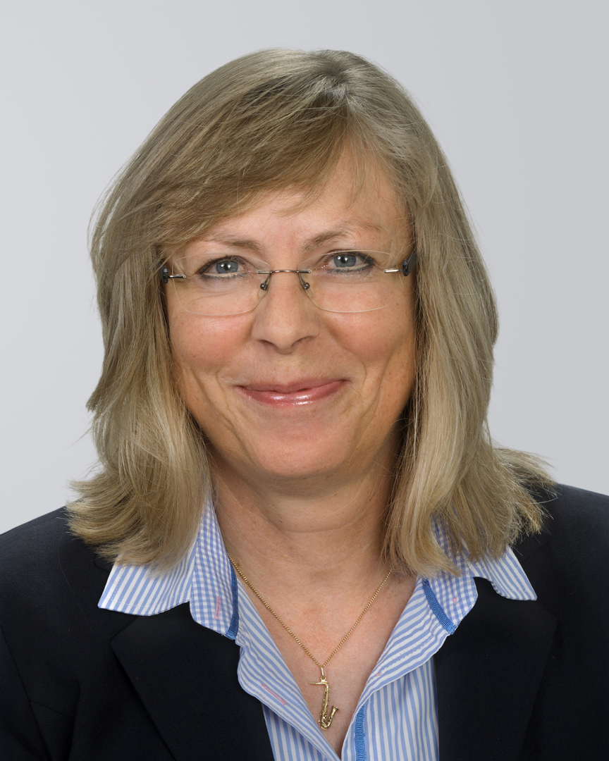 Frau mit blonden, schulterlangen Haaren, Brille und dunklem Blazer vor neutralem Hintergrund.