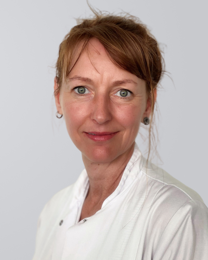 Frau mit kurzen, roten Haaren, blauen Augen und weißem Arztkittel vor neutralem Hintergrund.
