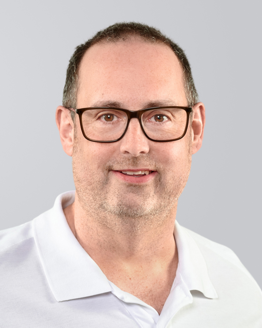 Mann mit Brille und kurzem Haar lächelt in einem weißen Hemd vor einem grauen Hintergrund.
