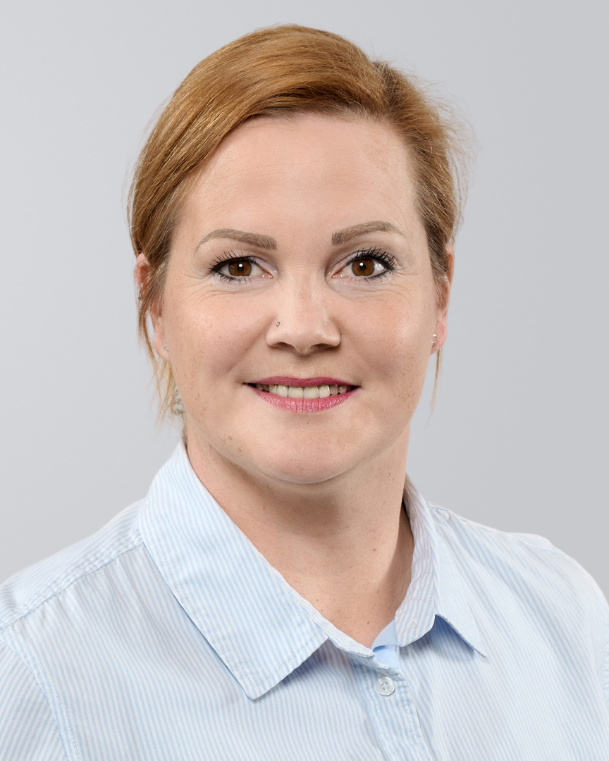 Porträt einer lächelnden Frau mit kurzen, roten Haaren und hellblauem Hemd vor einem grauen Hintergrund.