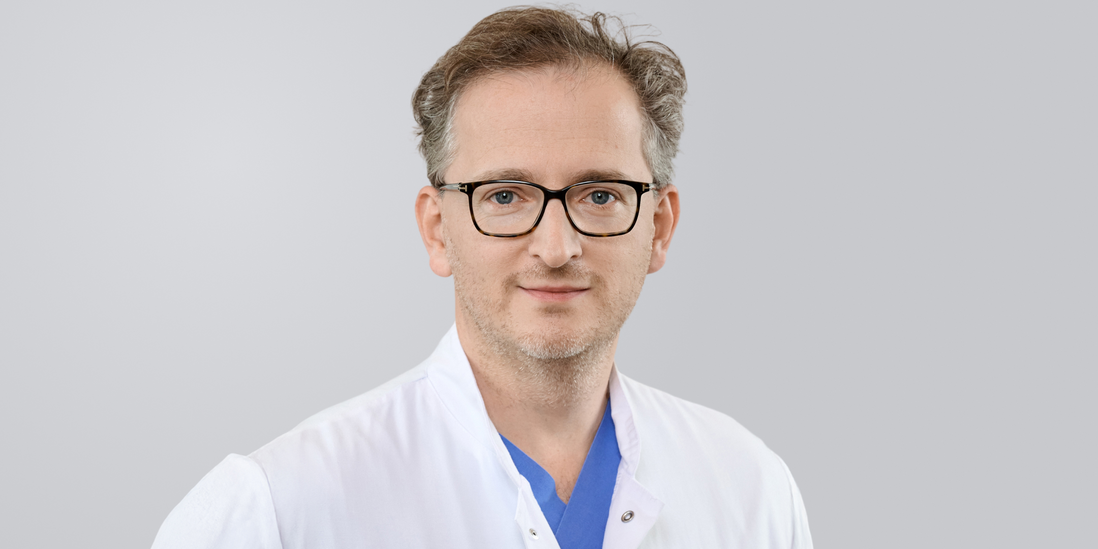 Ein Arzt mit grauen Haaren, Brille und weißem Kittel vor einem grauen Hintergrund.