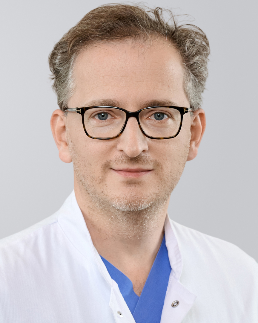 Ein Arzt mit grauen Haaren, Brille und weißem Kittel vor einem grauen Hintergrund.