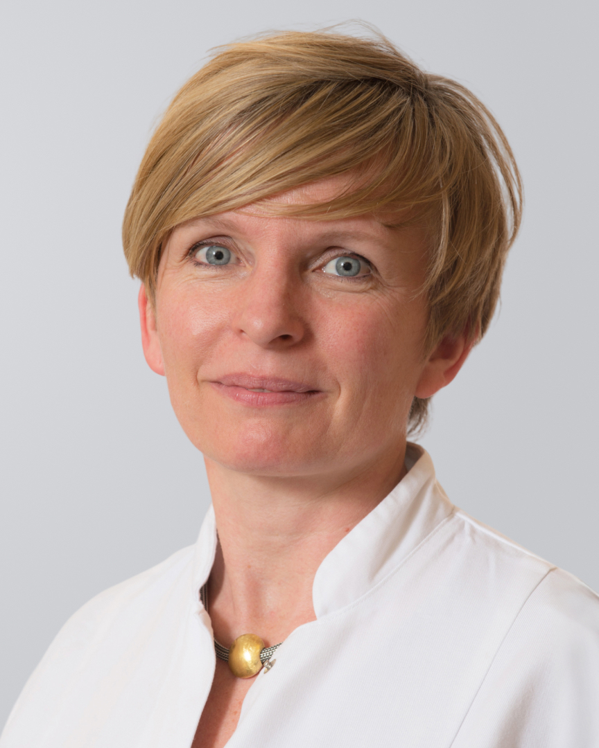 Porträt einer Frau mit kurzem, blonden Haar und weißem Kittel vor grauem Hintergrund.