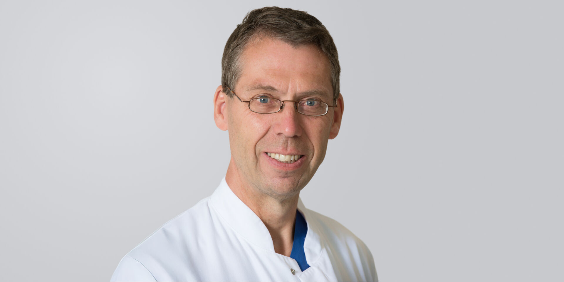 Lächelnder Mann mit weißem Arztkittel und Brille vor neutralem Hintergrund.
