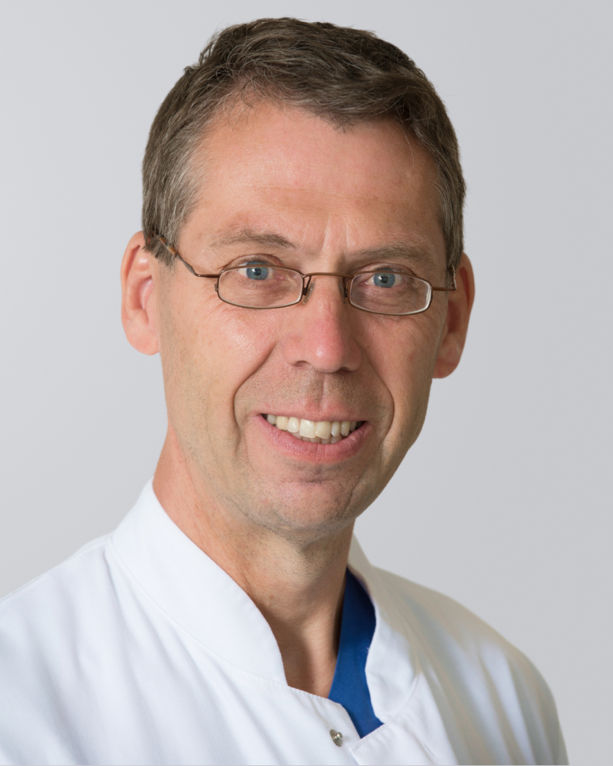 Lächelnder Mann mit weißem Arztkittel und Brille vor neutralem Hintergrund.