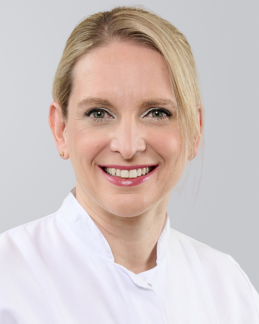 Lächelnde Frau mit blonden Haaren und weißem Arztkittel vor grauem Hintergrund.