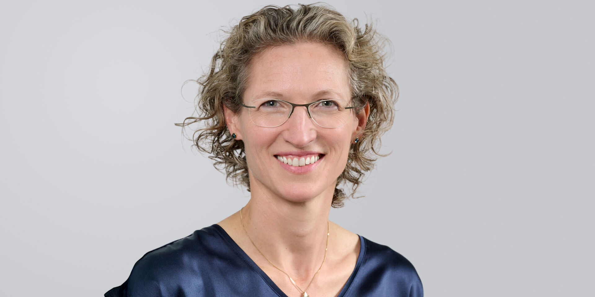 Porträt einer lächelnden Frau mit lockigem, blondem Haar und blauer Bluse vor einem grauen Hintergrund.