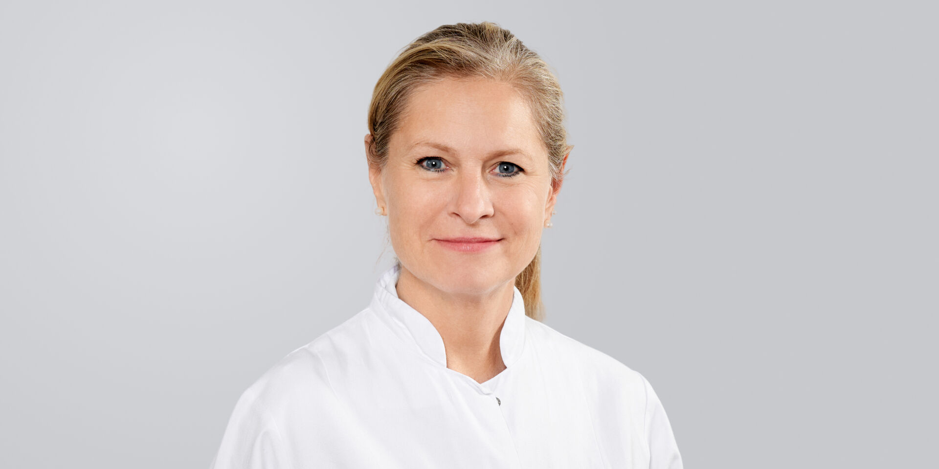 Blonde Frau in weißem Arztkittel lächelt vor grauem Hintergrund.