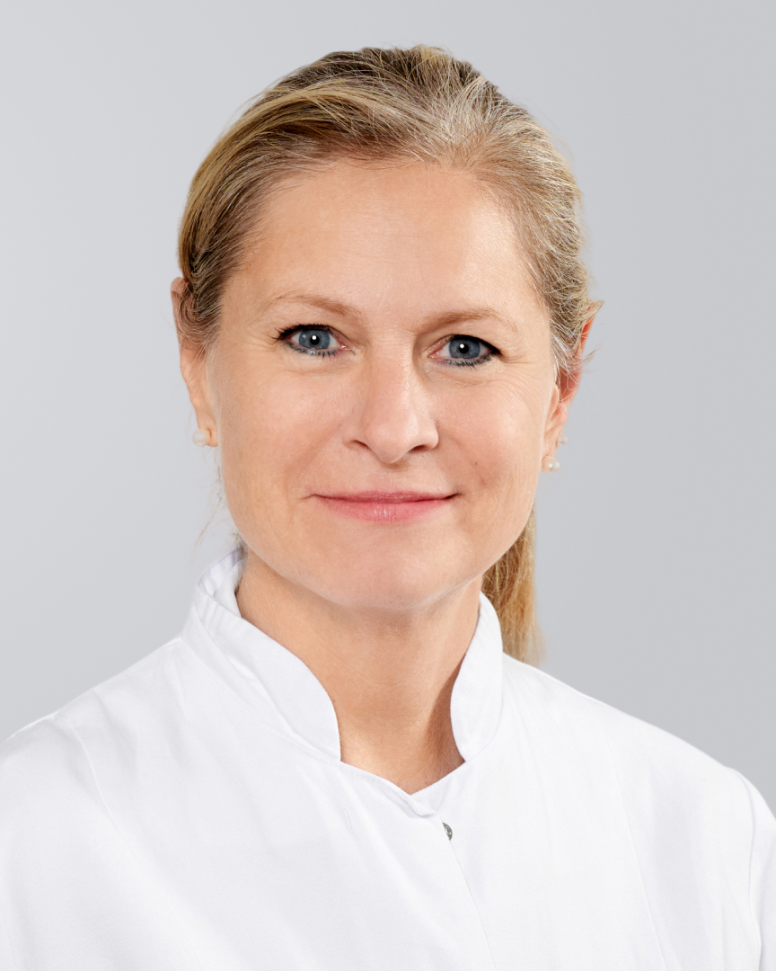 Blonde Frau in weißem Arztkittel lächelt vor grauem Hintergrund.