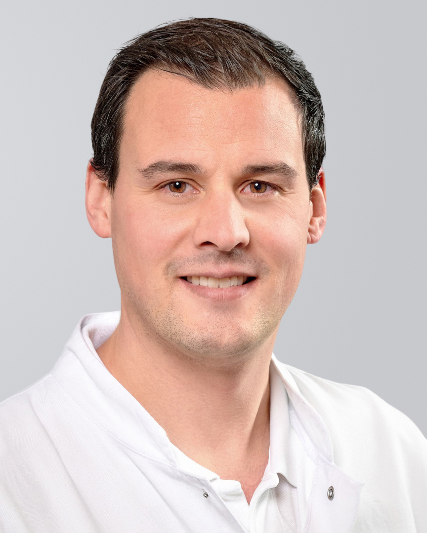 Lächelnder Mann mit kurzen, dunklen Haaren und weißem Kittel vor einem grauen Hintergrund.