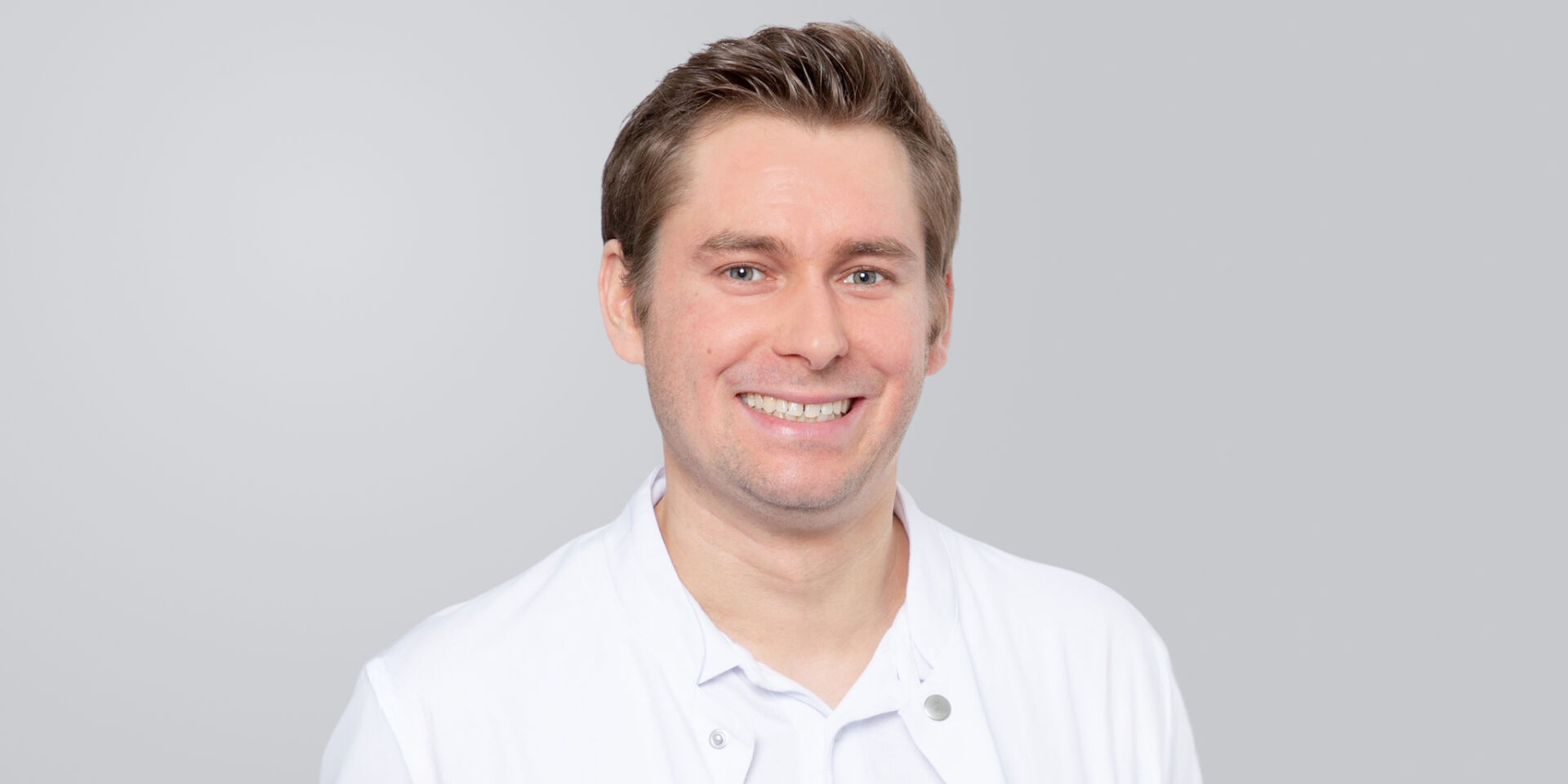 Lächelnder Mann mit kurzen Haaren und weißem Kittel vor grauem Hintergrund.