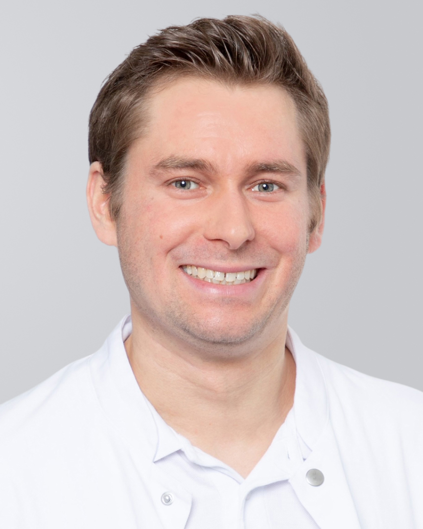 Lächelnder Mann mit kurzen Haaren und weißem Kittel vor grauem Hintergrund.