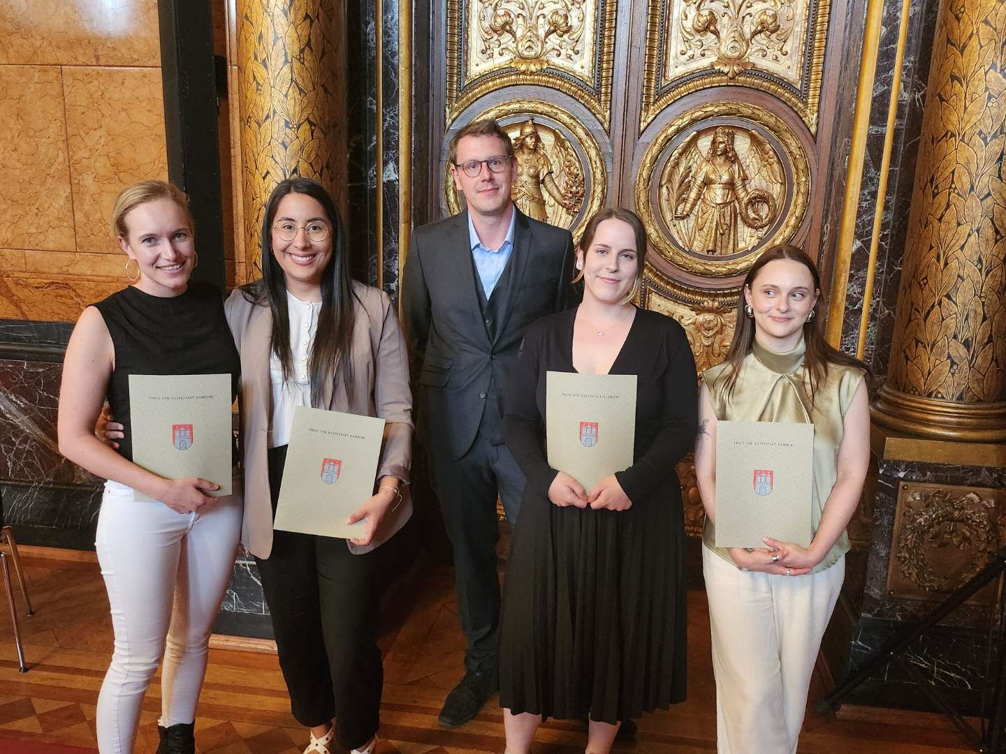 Fünf Menschen in förmlicher Kleidung stehen vor einer prunkvollen Goldtür. Jeweils zwei Frauen links und rechts des Mannes in der Bildmitte halten Zeugnisumschläge in die Kamera.