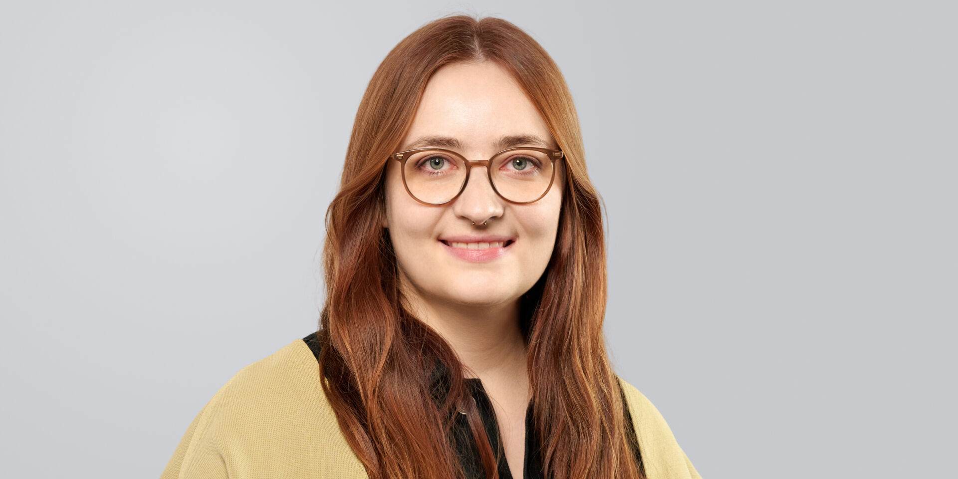 Porträt einer lächelnden Person mit langen, roten Haaren und Brille, die einen gelben Pullover trägt.