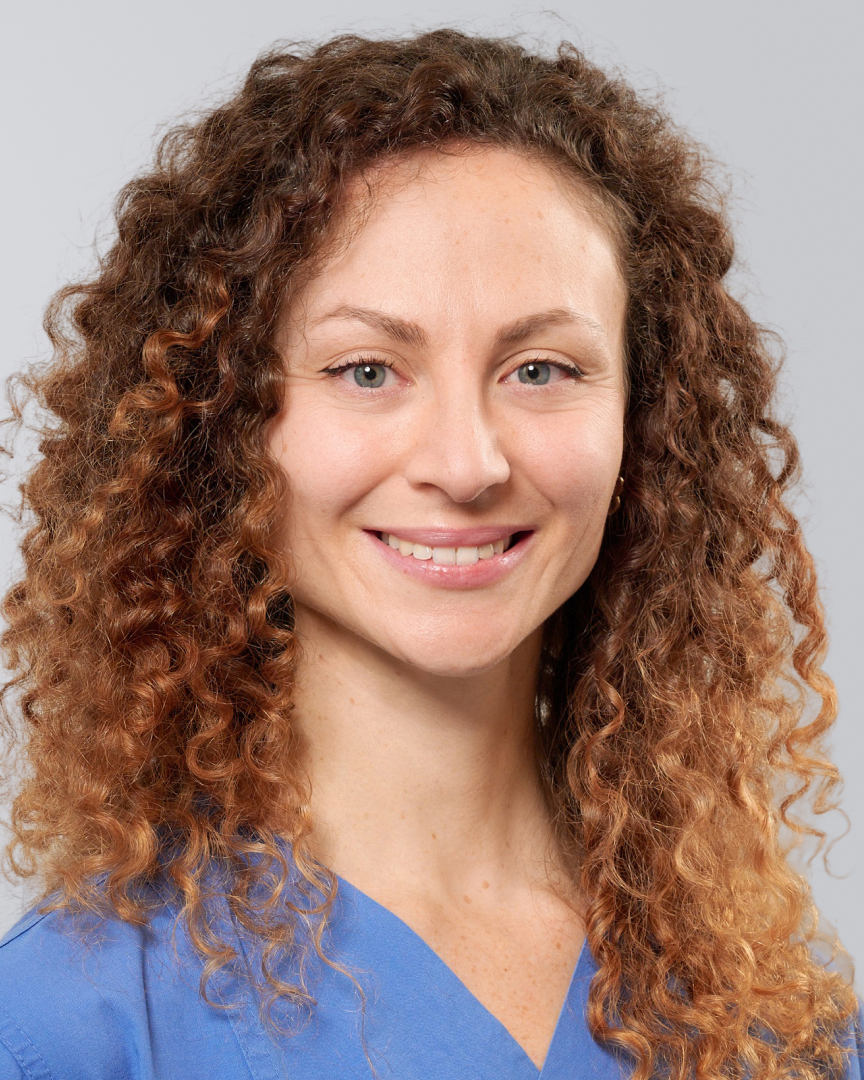 Frau mit lockigem, braunem Haar und blauer Arbeitskleidung vor einem grauen Hintergrund.