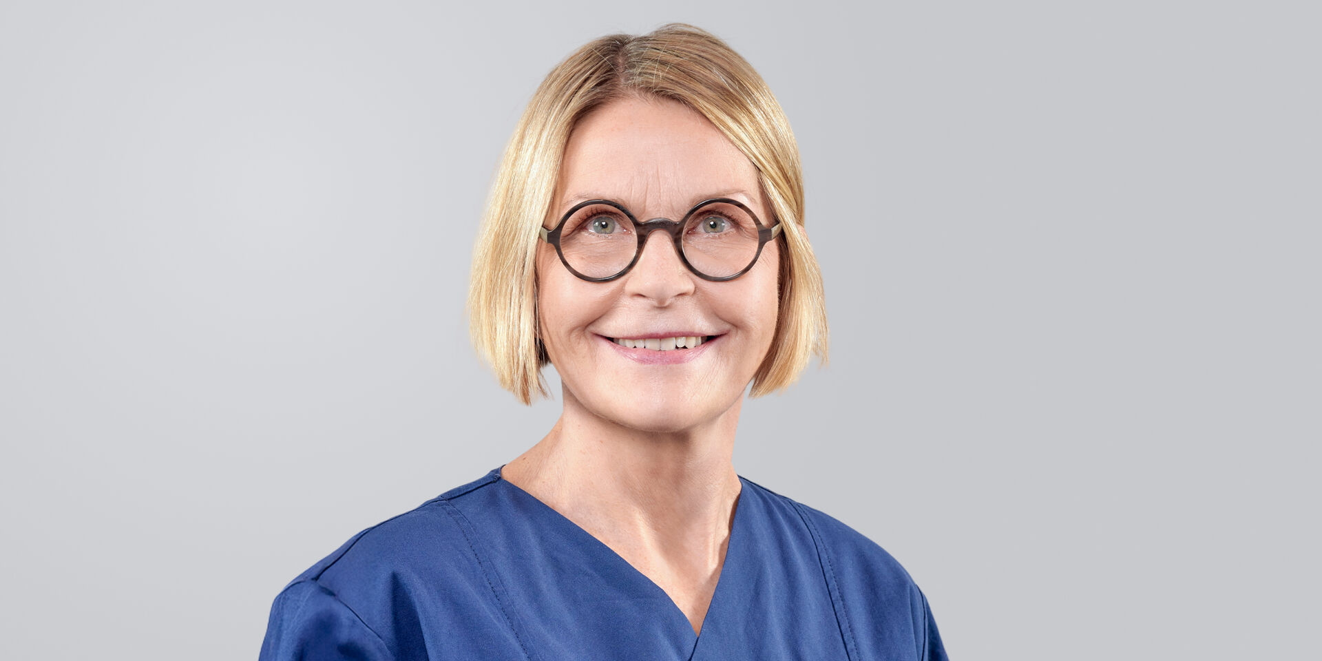 Lächelnde Frau mit kinnlangen blonden Haaren, Brille und blauer Arbeitskleidung vor neutralem Hintergrund.