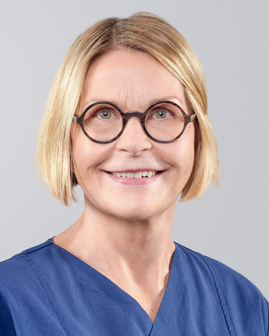 Lächelnde Frau mit kinnlangen blonden Haaren, Brille und blauer Arbeitskleidung vor neutralem Hintergrund.