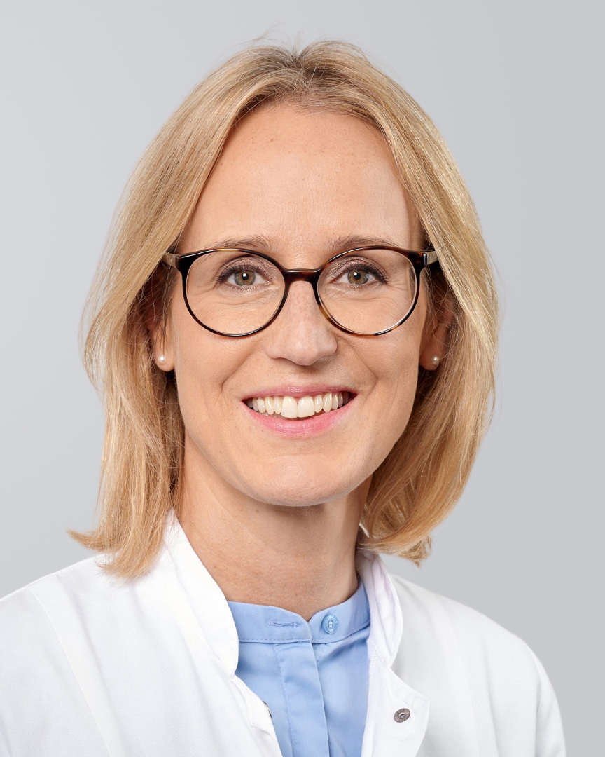 Lächelnde Frau mit Brille, blondem Haar und weißen Kittel vor einem grauen Hintergrund.