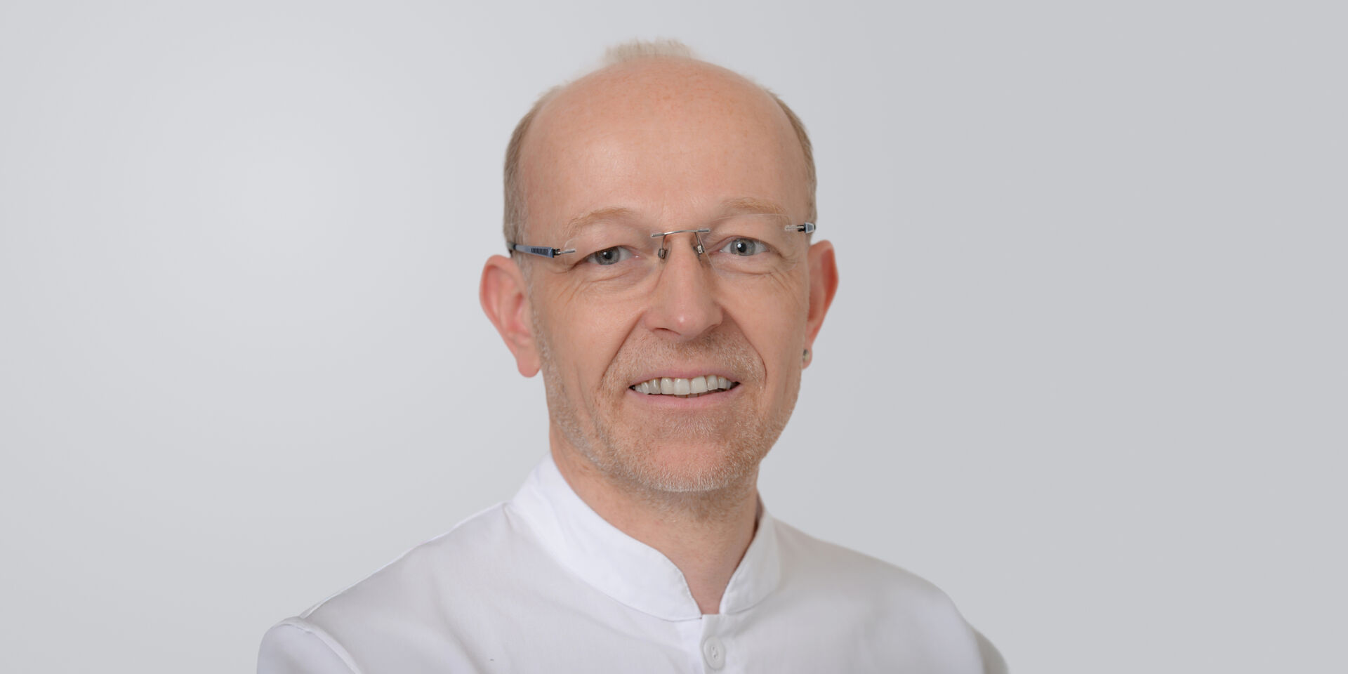 Ein lächelnder Mann mit Brille, grauem Haar Hemd und Arztkittel vor einem neutralen Hintergrund.