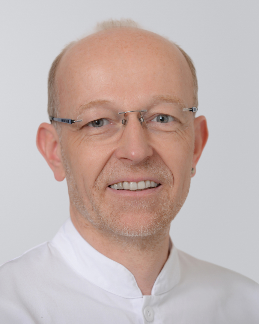 Ein lächelnder Mann mit Brille, grauem Haar Hemd und Arztkittel vor einem neutralen Hintergrund.