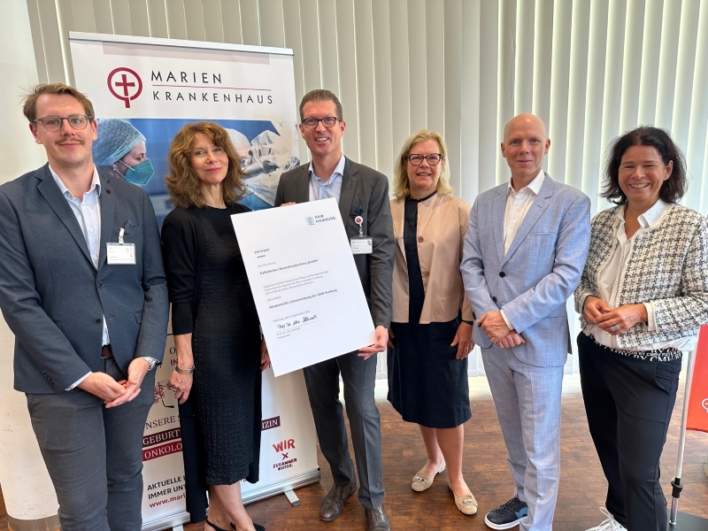 Eine Gruppe Menschen steht vor einer Stele des Marienkrankenhauses. Ein Mann in der Mitte hält eine übergroße Urkunde in die Kamera.