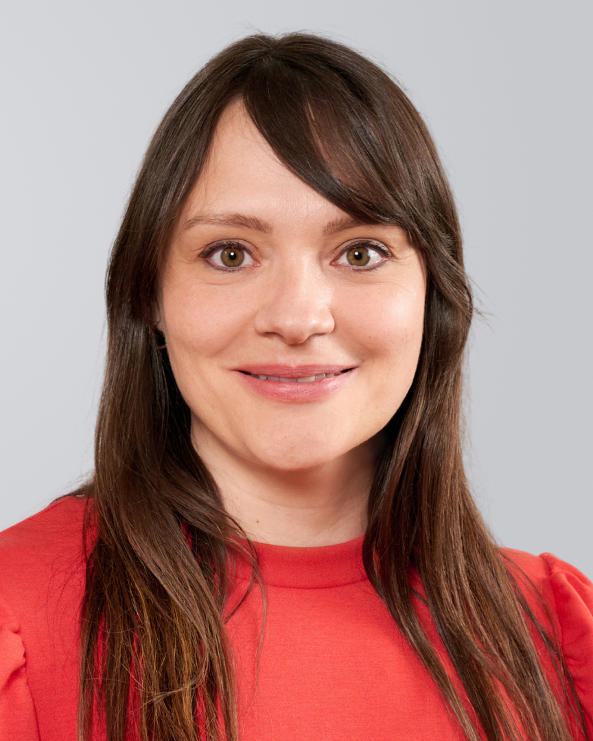 Lächelnde Frau mit langen, braunen Haaren und rotem Oberteil vor einem grauen Hintergrund.