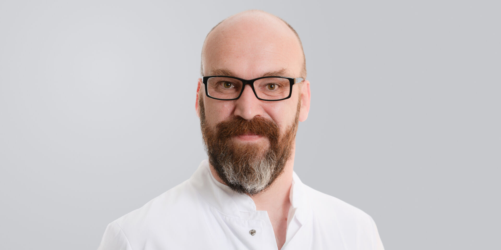 Lächelnder Mann mit Brille, Bart und weißem Kittel vor einem grauen Hintergrund.