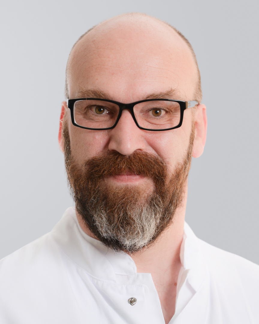 Lächelnder Mann mit Brille, Bart und weißem Kittel vor einem grauen Hintergrund.
