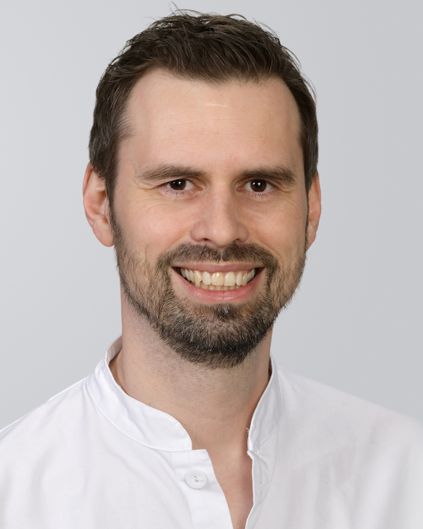 Lächelnder Mann mit dunklem Haar, Bart und weißem Hemd vor neutralem Hintergrund.