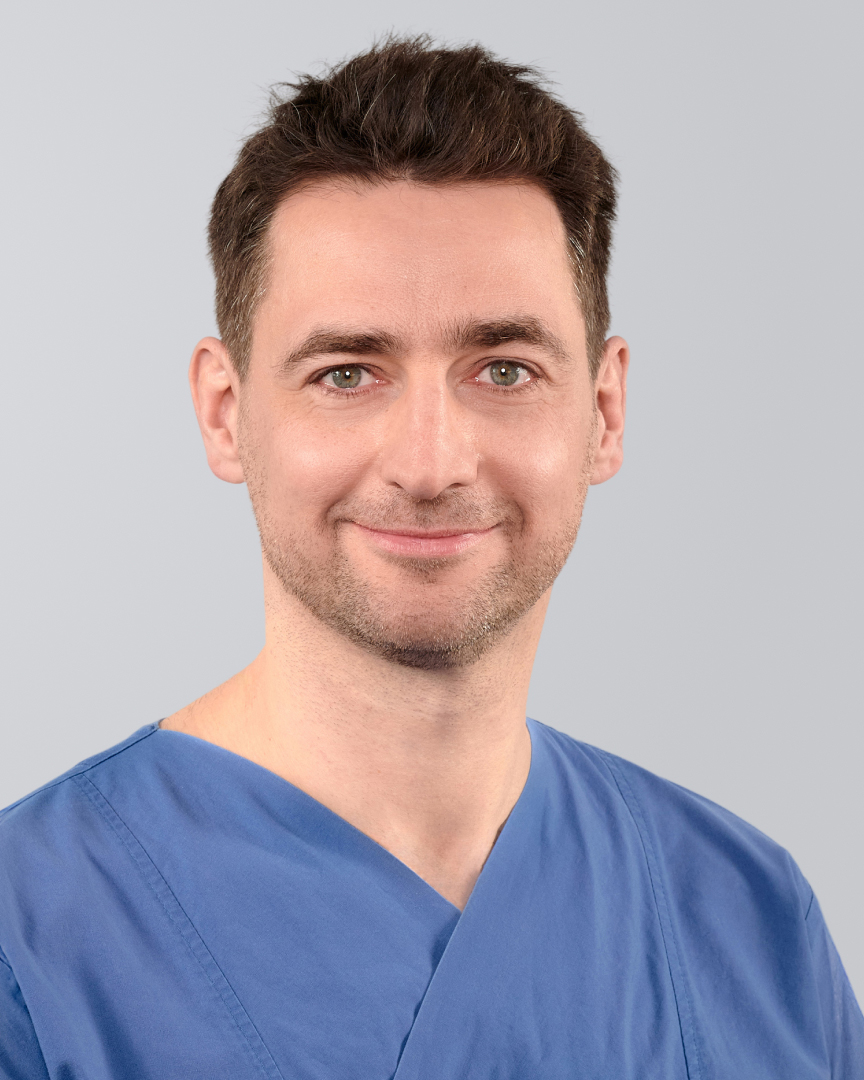 Mann in blauer medizinischer Kleidung mit kurzem, dunklem Haar und lächelndem Gesicht vor neutralem Hintergrund.