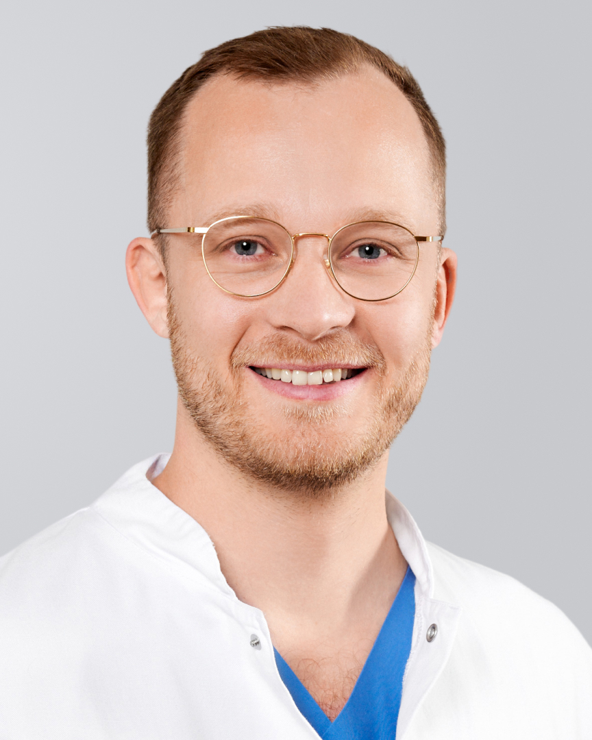 Lächelnder Mann mit kurzen Haaren, Brille und weißem Kittel vor neutralem Hintergrund.