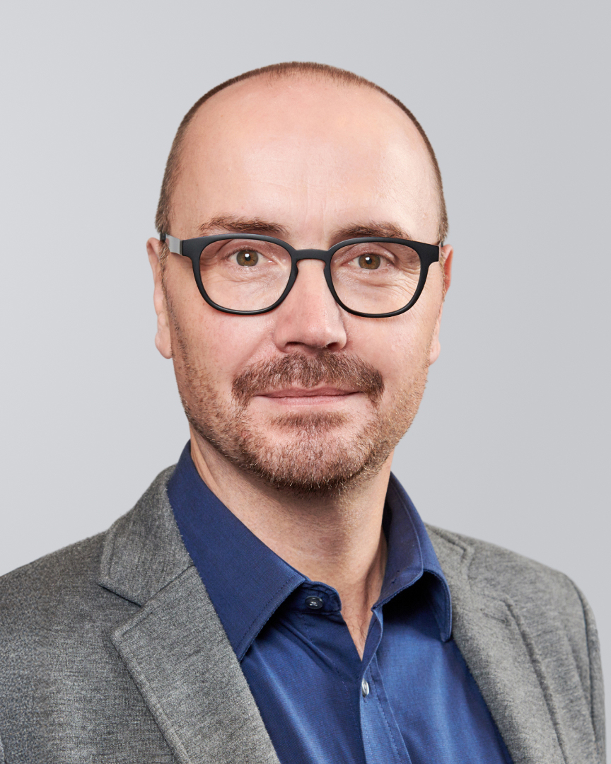 Porträt eines Mannes mit Brille, kurzen Haaren und grauem Sakko vor neutralem Hintergrund.