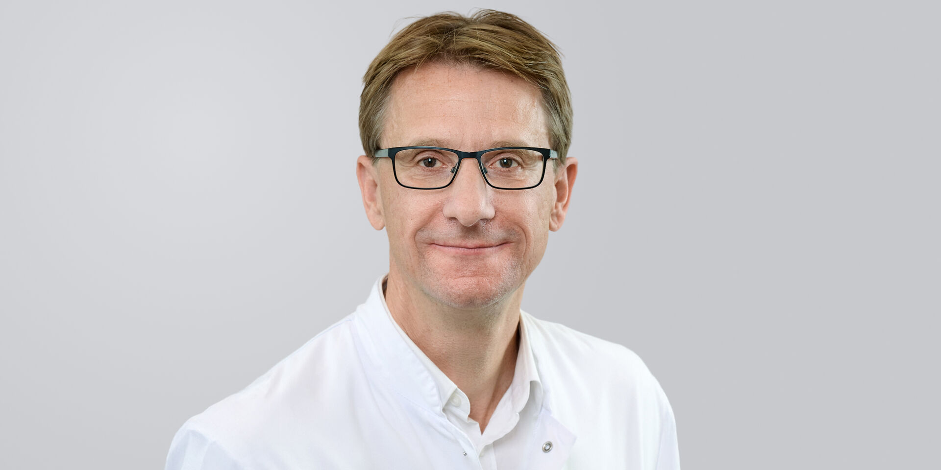 Porträt eines lächelnden Mannes mit Brille und weißem Kittel vor einem neutralen Hintergrund.