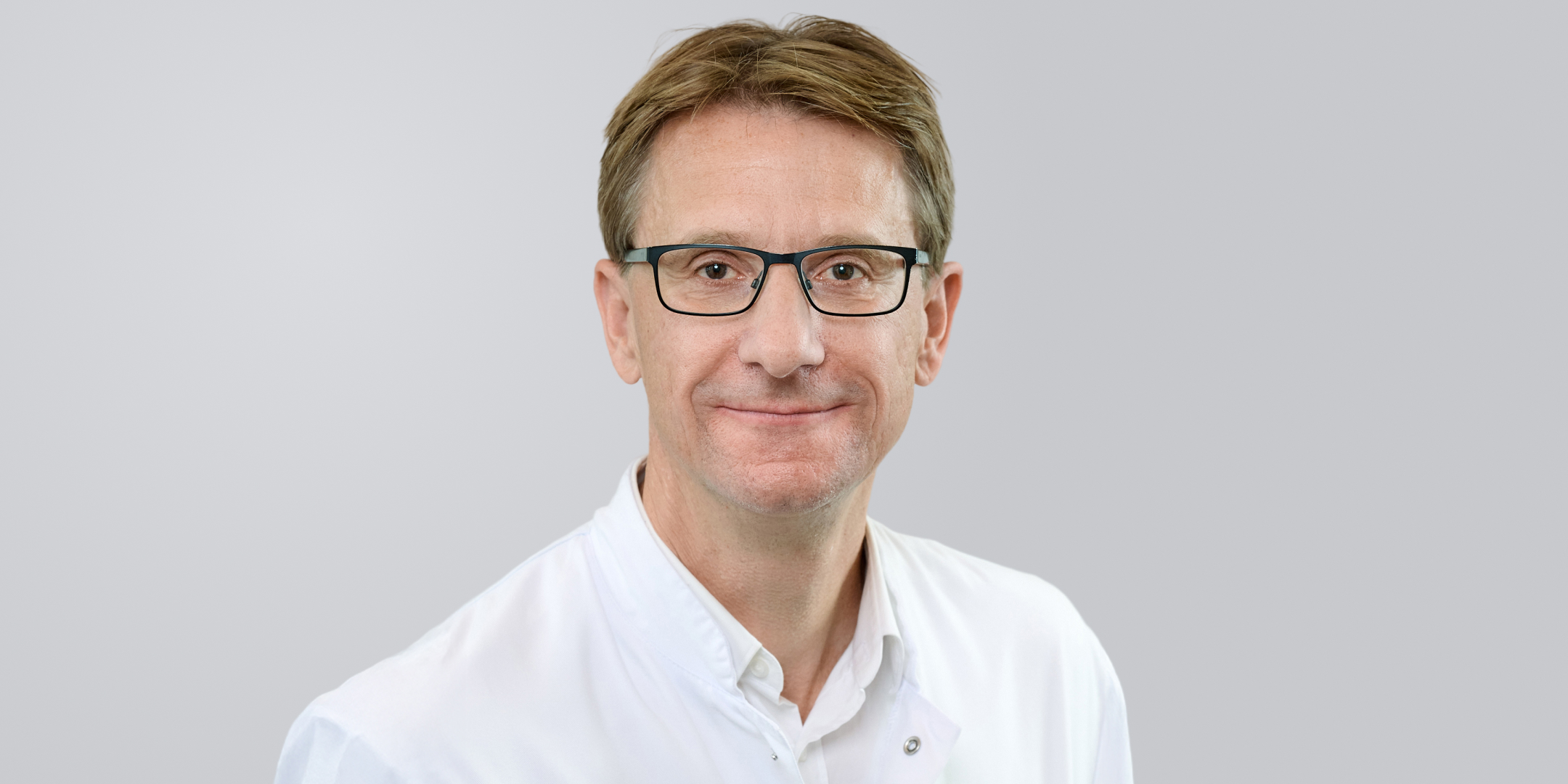 Porträt eines lächelnden Mannes mit Brille und weißem Kittel vor einem neutralen Hintergrund.