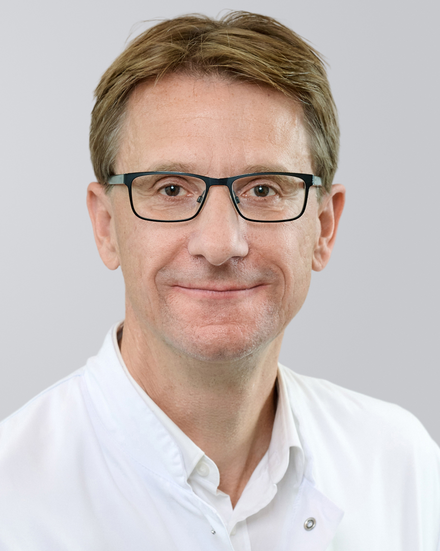 Porträt eines lächelnden Mannes mit Brille und weißem Kittel vor einem neutralen Hintergrund.