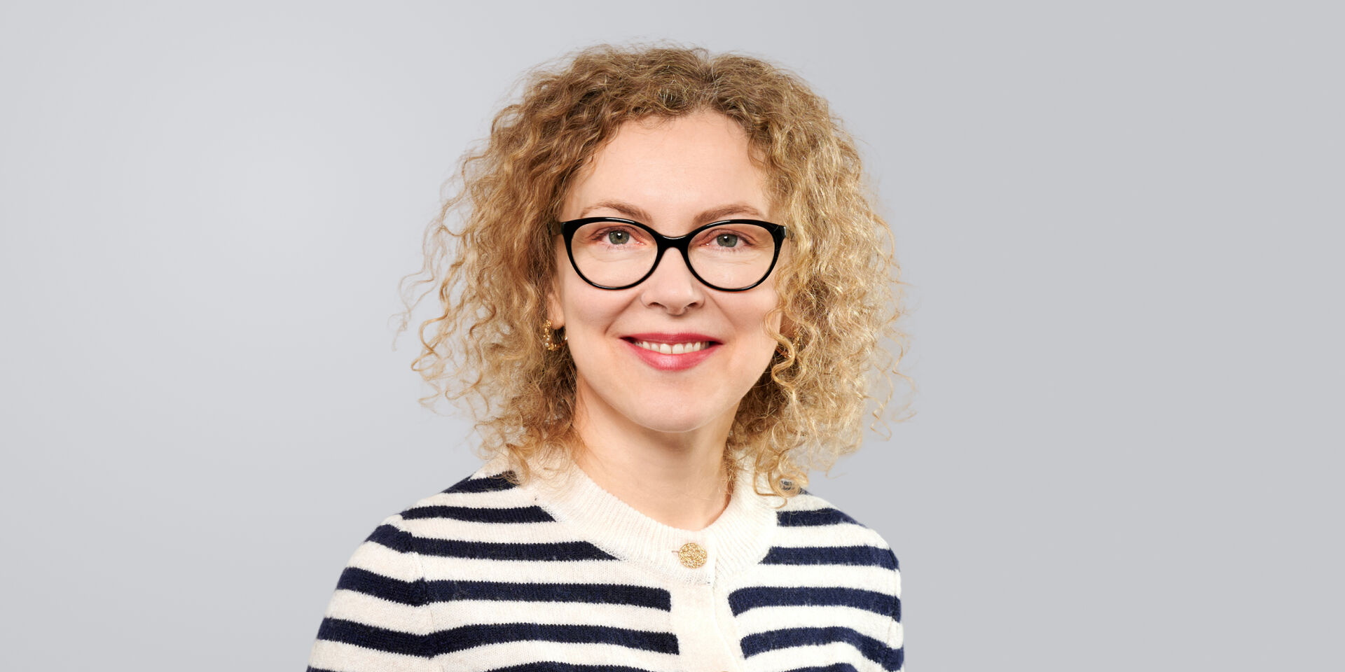 Frau mit lockigem, blondem Haar, Brille und gestreiftem Oberteil vor grauem Hintergrund.