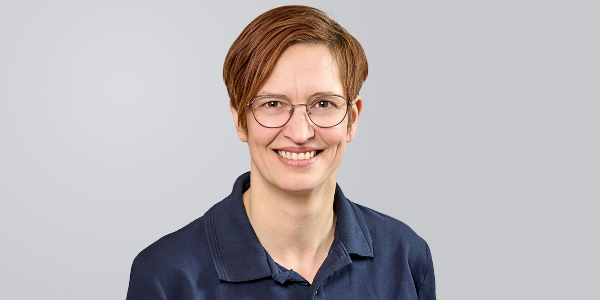 Frau mit kurzen, braunen Haaren, Brille und blauem Oberteil vor einem grauen Hintergrund.