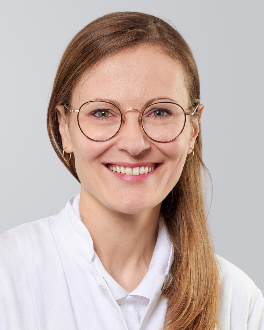 Lächelnde Frau mit langen, blonden Haaren, Brille und weißem Arztkittel vor einem grauen Hintergrund.