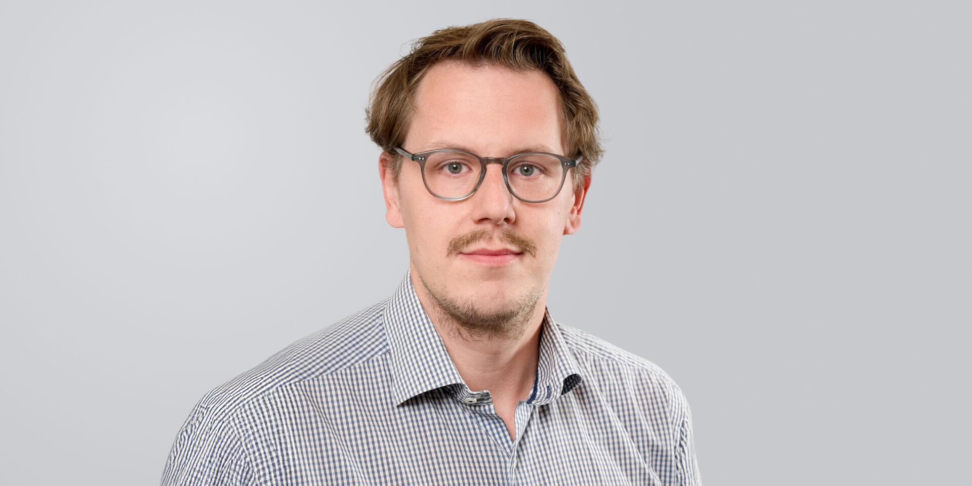 Mann mit Brille und kariertem Hemd vor neutralem Hintergrund.
