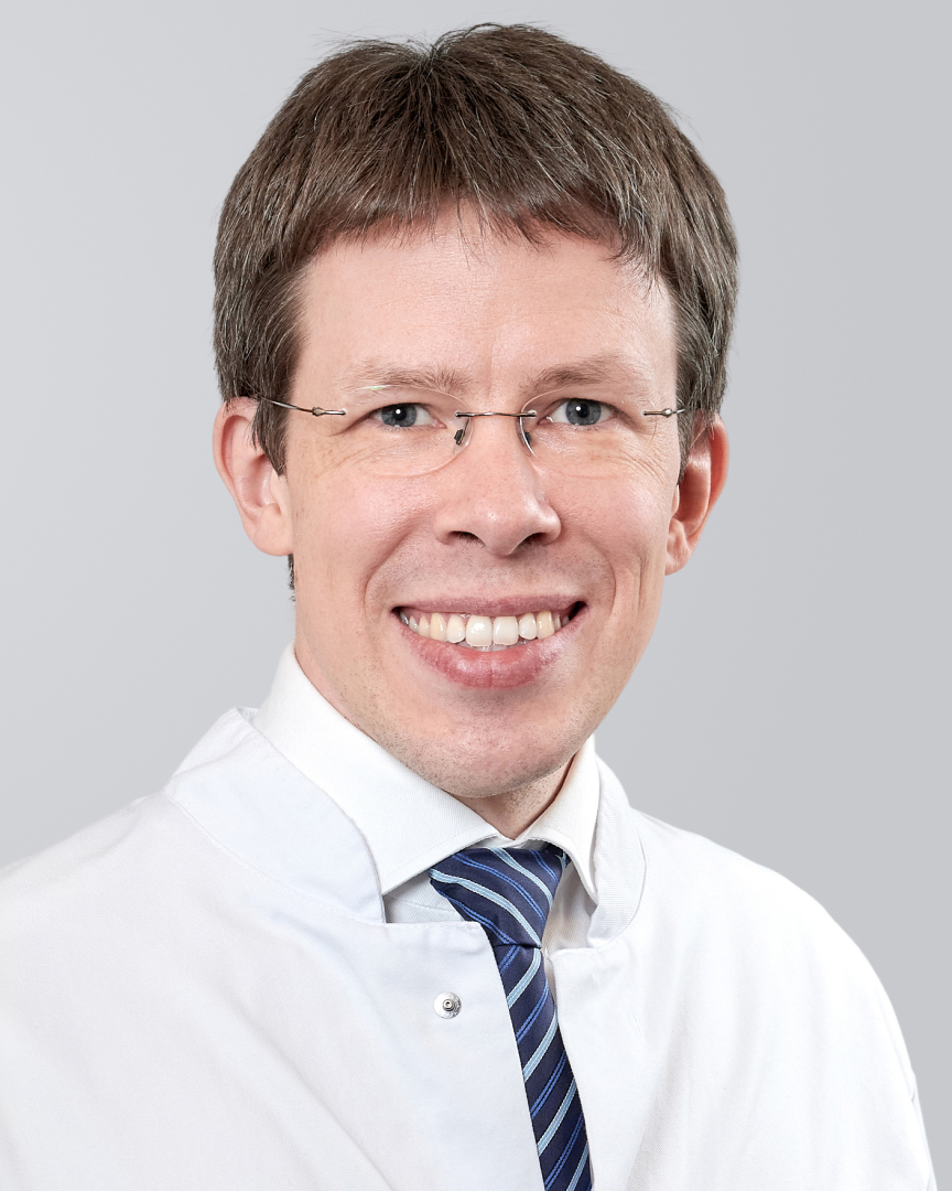Lächelnder Mann mit Brille, einem weißen Hemd und Krawatte vor einem grauen Hintergrund.