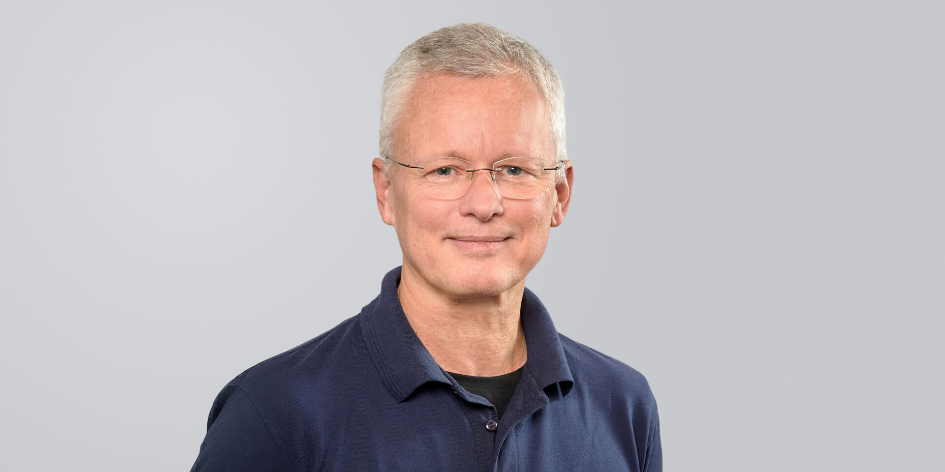 Mann mit grauen Haaren und Brille, lächelt in einem dunkelblauen Hemd vor einem neutralen Hintergrund.