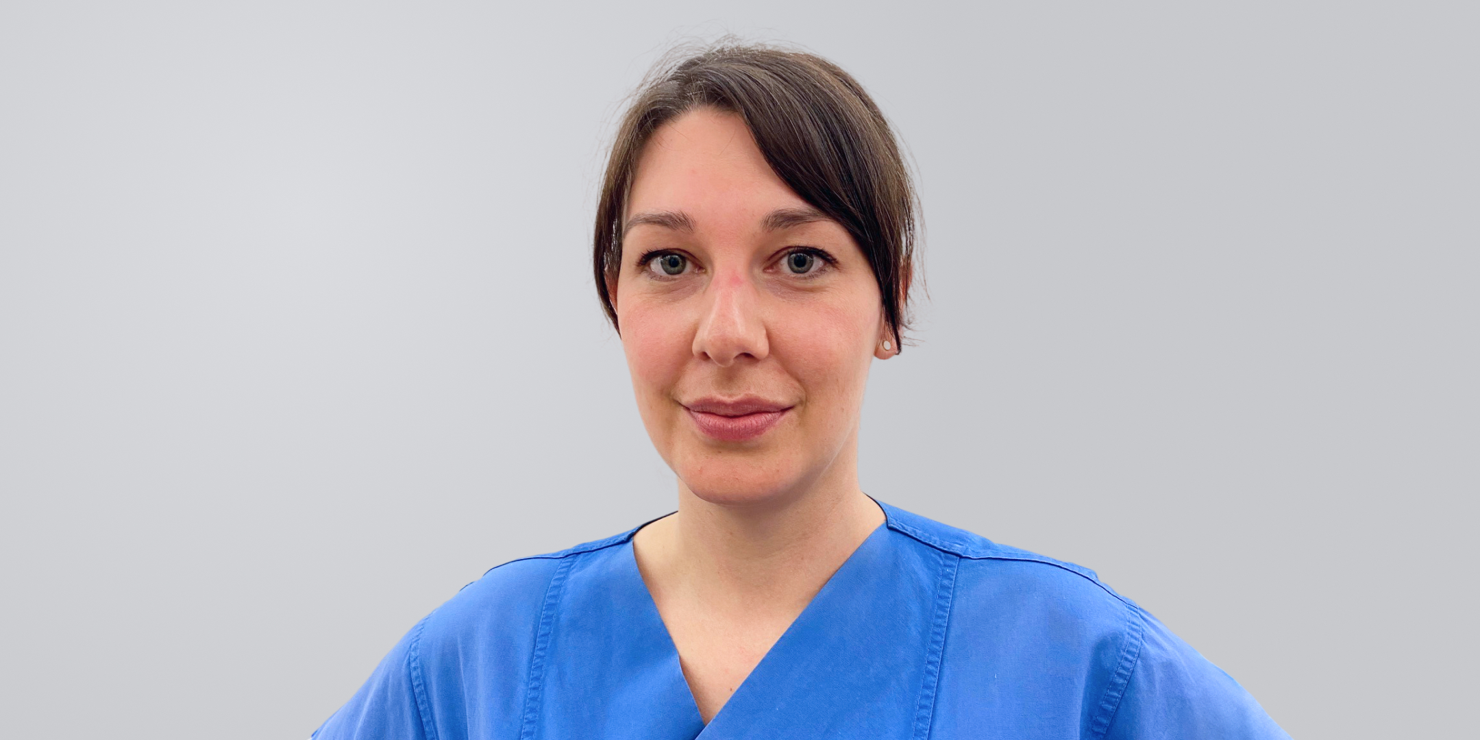 Frau mit dunklem Haar und blauer medizinischer Kleidung vor einem neutralen Hintergrund.